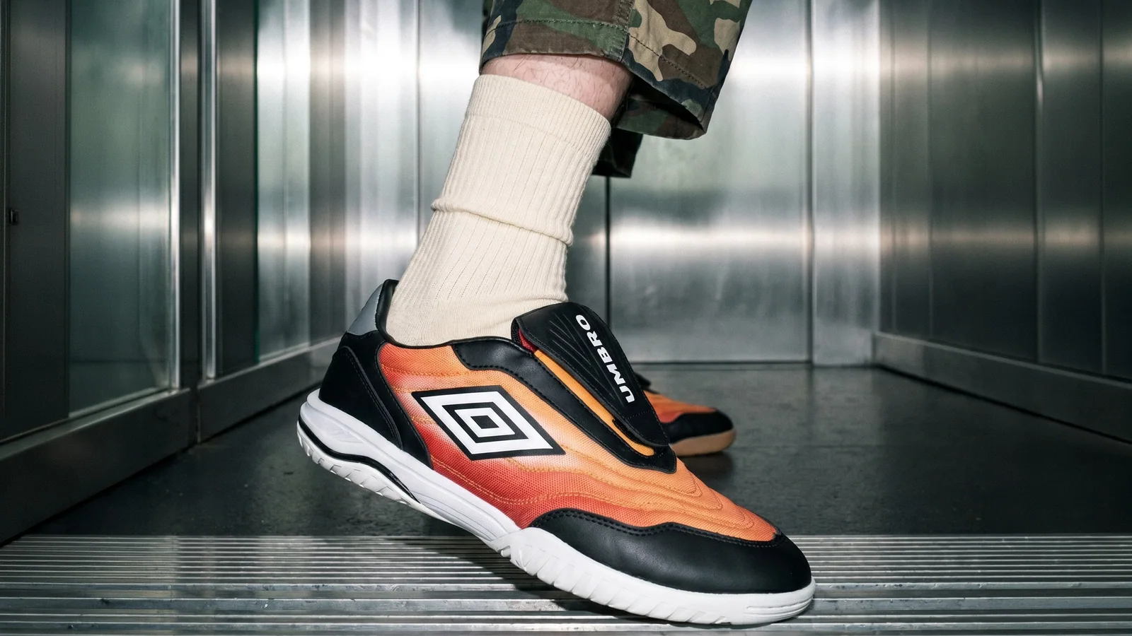Orange Umbro footwear styled with cream socks and camouflage shorts inside a metal elevator